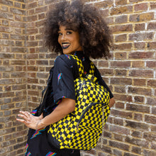 Load image into Gallery viewer, Cindy is stood outside in front of a brick wall wearing the CHOK yellow checker vegan backpack on her back whilst looking over her shoulder to camera. The backpack is yellow and black checkerboard all over print with two front zip pockets, two side pockets and a silver decorative chain going across the front.