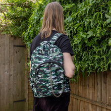 Load image into Gallery viewer, Jack is stood in a garden area modelling the classic camouflage camo vegan backpack. The bag is facing towards the camera to highlight the front camo print, two front zip pockets, two elastic side pockets, detachable silver chain and top handle.
