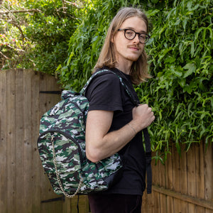 Jack is stood in a garden area modelling the classic camouflage camo vegan backpack. The bag is facing towards the camera to highlight the front camo print, two front zip pockets, two elastic side pockets, detachable silver chain and top handle.