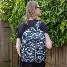 Load image into Gallery viewer, Jack is stood in a garden area modelling the forest camouflage camo vegan backpack. The bag is facing towards the camera to highlight the front camo print, two front zip pockets, two elastic side pockets, detachable silver chain and top handle.