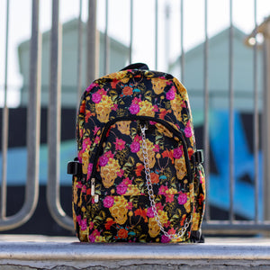 The floral gold skull nylon chain vegan backpack sat on a skatepark ramp. The bag is facing forward to highlight the flowers and skulls print, two zipped pockets, two elastic side pockets and detachable decorative silver chain. 