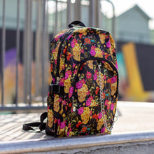 Load image into Gallery viewer, The floral gold skull nylon chain vegan backpack sat on a skatepark ramp. The bag is facing forward angled right to highlight the flowers and skulls print, two zipped pockets, two elastic side pockets and detachable decorative silver chain. 
