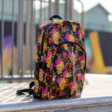 Load image into Gallery viewer, The floral gold skull nylon chain vegan backpack sat on a skatepark ramp. The bag is facing forward angled right to highlight the flowers and skulls print, two zipped pockets, two elastic side pockets and detachable decorative silver chain.