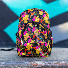 Load image into Gallery viewer, The floral gold skull nylon chain vegan backpack sat on a skatepark bench. The bag is facing forward to highlight the flowers and skulls print, two zipped pockets, two elastic side pockets and detachable decorative silver chain.