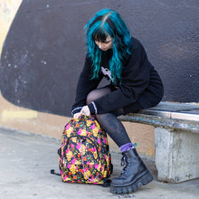 Load image into Gallery viewer, Model sat on a skatepark bench adjusting the floral gold skull nylon chain vegan backpack. The bag is facing forward to highlight the flowers and skulls print, two zipped pockets, two elastic side pockets and detachable decorative silver chain.
