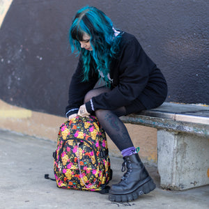 Model sat on a skatepark bench adjusting the floral gold skull nylon chain vegan backpack. The bag is facing forward to highlight the flowers and skulls print, two zipped pockets, two elastic side pockets and detachable decorative silver chain.