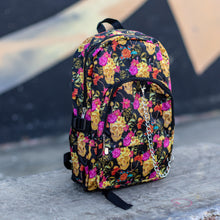 Load image into Gallery viewer, The floral gold skull nylon chain vegan backpack sat on a skatepark bench. The bag is facing forward angled right to highlight the flowers and skulls print, two zipped pockets, two elastic side pockets and detachable decorative silver chain. 

