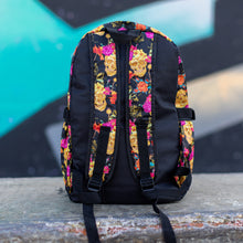 Load image into Gallery viewer, The floral gold skull nylon chain vegan backpack sat on a skatepark bench. The bag is facing away from the camera to highlight the plain black bag, two elastic side pockets and two adjustable padded shoulder straps.