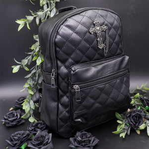 The GothX quilted cross vegan mini backpack on a black studio background with black roses and leaves surrounding it. Quilted front detailing with a studded cross with hanging chain appliqué and zip front pocket. Black vegan leather with gunmetal grey detailing. The bag is angled forward to the right to show off the depth of the bag.