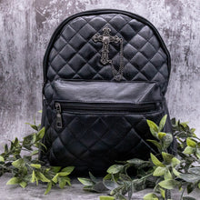 Load image into Gallery viewer, The GothX quilted cross vegan mini backpack on a grey studio background with leaves surrounding it. Quilted front detailing with a studded cross with hanging chain appliqué and zip front pocket. Black vegan leather with gunmetal grey detailing. The bag is facing forward.
