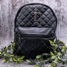 Load image into Gallery viewer, The GothX quilted cross vegan mini backpack on a grey studio background with leaves surrounding it. Quilted front detailing with a studded cross with hanging chain appliqué and zip front pocket. Black vegan leather with gunmetal grey detailing. The bag is facing forward.