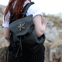 Load image into Gallery viewer, GothX Skull and Star Black Vegan Tassel Tie Backpack shown being held by a model wearing pin stripe dungarees and a white short sleeve top. They are walking away from the camera with the bag on one shoulder whilst their hand rests on the front strap. Bag has adjustable vegan leather straps with tassel tie and metal magnetic clip close with large zipped front pocket.