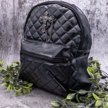 Load image into Gallery viewer, The GothX quilted cross vegan mini backpack on a grey studio background with leaves surrounding it. Quilted front detailing with a studded cross with hanging chain appliqué and zip front pocket. Black vegan leather with gunmetal grey detailing. The bag is angled to the left to show the depth of the bag.