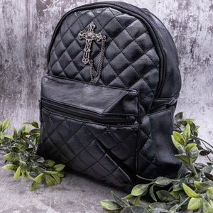 The GothX quilted cross vegan mini backpack on a grey studio background with leaves surrounding it. Quilted front detailing with a studded cross with hanging chain appliqué and zip front pocket. Black vegan leather with gunmetal grey detailing. The bag is angled to the left to show the depth of the bag. 