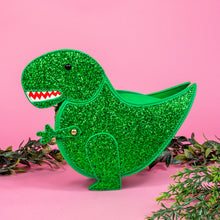 Load image into Gallery viewer, The kawaii green glitter dino vegan bag on a pink studio background with green foliage. The bag is facing forward to highlight the green glittered side, dinosaur face, gold metal detailing, detachable strap and moveable arm.