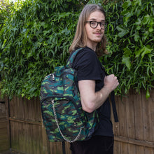 Load image into Gallery viewer, Jack is stood in a garden area modelling the jungle camouflage camo vegan backpack. The bag is facing towards the camera to highlight the front camo print, two front zip pockets, two elastic side pockets, detachable silver chain and top handle.