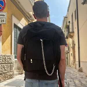 Model is stood in amongst Spanish yellow buildings wearing the black nylon vegan backpack with chain on their back. They are facing away from the camera with both hands by their side. The backpack is all black with two front zip pockets, two side pockets and a silver decorative chain across the front to the side.