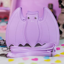 Load image into Gallery viewer, The GothX LIMITED EDITION Pastel Lilac Purple Bat Vegan Shoulder Bag on a pastel purple background with multicoloured confetti, pastel pink coffin shelving and black skull bottles surrounding it. The bag is facing forward to highlight the embroidered detailing, crystal eyes and detachable adjustable strap.
