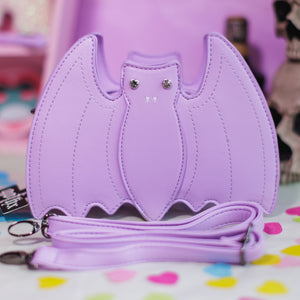 The GothX LIMITED EDITION Pastel Lilac Purple Bat Vegan Shoulder Bag on a pastel purple background with multicoloured confetti, pastel pink coffin shelving and black skull bottles surrounding it. The bag is facing forward to highlight the embroidered detailing, crystal eyes and detachable adjustable strap.