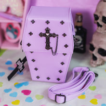Load image into Gallery viewer, The GothX Pastel Lilac Mini Coffin Vegan Cross Body Bag on a pastel purple background with pastel pink coffin shelving, skulls and black faux skull poison bottles in the background. The bag is facing forward to highlight the cross studs, cross & chain centrepiece, detachable chain and detachable adjustable shoulder strap.