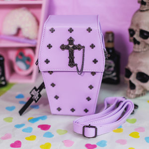The GothX Pastel Lilac Mini Coffin Vegan Cross Body Bag on a pastel purple background with pastel pink coffin shelving, skulls and black faux skull poison bottles in the background. The bag is facing forward to highlight the cross studs, cross & chain centrepiece, detachable chain and detachable adjustable shoulder strap.