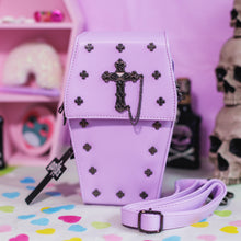 Load image into Gallery viewer, The GothX Pastel Lilac Mini Coffin Vegan Cross Body Bag on a pastel purple background with pastel pink coffin shelving, skulls and black faux skull poison bottles in the background. The bag is facing forward to highlight the cross studs, cross & chain centrepiece, detachable chain and detachable adjustable shoulder strap.