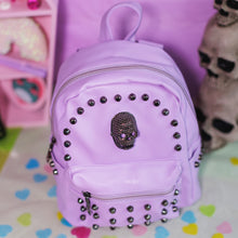 Load image into Gallery viewer, The GothX Pastel Lilac Skull Head Small Studs Vegan Mini Backpack on a pastel purple background with skulls and pastel pink coffin shelf in the background. The bag is facing forward angled right to highlight the dark grey metal stud detailing, diamanté effect skull, main zip compartment, front zip pocket, two side slip pockets and top handle.