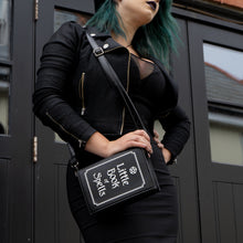 Load image into Gallery viewer, Model wearing an all black goth style outfit holding the gothx little book of spells vegan shoulder bag on one shoulder. The witchy inspired vegan leather bag is facing towards the camera to highlight the white printed design of a pentagram, framing and text reading little book of spells.