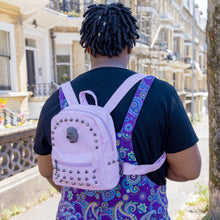 Load image into Gallery viewer, The GothX Pastel Pink Skull Head Small Studs Vegan Mini Backpack being worn by an alternative model wearing a black tshirt and the run and fly purple paisley pinafore and the mini backpack on their back. They are facing away to show the front of the bag highlighting the dark grey metal stud detailing, diamanté effect skull, front zip pocket, two side slip pockets and top handle.
