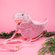 Load image into Gallery viewer, The kawaii pink glitter dino vegan bag on a pink studio background with green foliage. The vegan leather sparkly bag is facing forward to highlight the pastel pink glittered side, dinosaur face, gold metal detailing, detachable strap and moveable arm.