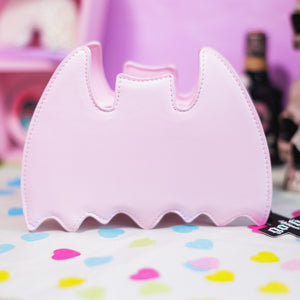 The GothX Pastel Pink Bat Vegan Shoulder Bag on a pastel purple background with multicoloured confetti, pastel pink coffin shelving and black skull bottles surrounding it. The bag is facing away to highlight the plain pastel pink back.
