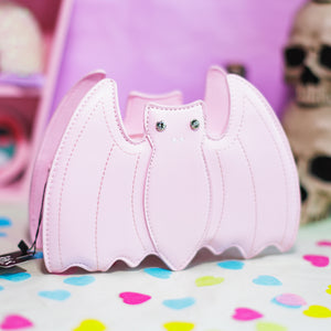 The GothX Pastel Pink Bat Vegan Shoulder Bag on a pastel purple background with multicoloured confetti, pastel pink coffin shelving and black skull bottles surrounding it. The bag is facing forward to highlight the embroidered detailing and crystal eyes.
