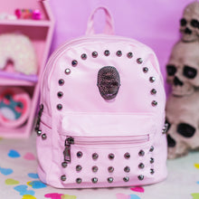 Load image into Gallery viewer, The GothX Pastel Pink Skull Head Small Studs Vegan Mini Backpack on a pastel purple background with skulls and pastel pink coffin shelf in the background. The vegan leather bag is facing forward to highlight the dark grey metal stud detailing, diamanté effect skull, front zip pocket, two side slip pockets and top handle.
