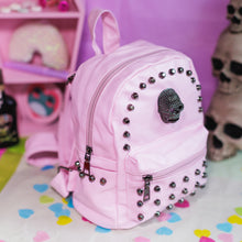Load image into Gallery viewer, The GothX Pastel Pink Skull Head Small Studs Vegan Mini Backpack on a pastel purple background with skulls and pastel pink coffin shelf in the background. The vegan leather bag is facing forward angled right to highlight the dark grey metal stud detailing, diamanté effect skull, main zip compartment, front zip pocket, two side slip pockets and top handle.