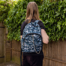 Load image into Gallery viewer, Jack is stood in a garden area modelling the pixel camouflage camo vegan backpack. The bag is facing towards the camera to highlight the front camo print, two front zip pockets, two elastic side pockets, detachable silver chain and top handle.