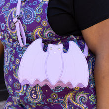 Load image into Gallery viewer, The GothX Pastel Lilac Purple Bat Vegan Shoulder Bag being modelled on a shoulder of someone wearing Run & Fly purple paisley pinafore. The vegan leather pastel bag is facing forward to highlight the embroidered detailing and crystal eyes.