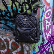 Load image into Gallery viewer, The GothX quilted cross vegan mini backpack sat outside on an alternative style colourful graffiti wall. The vegan leather bag is sat facing forward to highlight the quilted stitched front with a silver metal cross chain centre emblem, the front zip pocket, two side slip pockets, main top zip compartment and top handle.