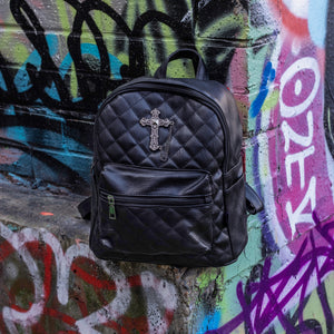 The GothX quilted cross vegan mini backpack sat outside on an alternative style colourful graffiti wall. The vegan leather bag is sat facing forward to highlight the quilted stitched front with a silver metal cross chain centre emblem, the front zip pocket, two side slip pockets, main top zip compartment and top handle.