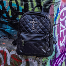Load image into Gallery viewer, The GothX quilted cross vegan mini backpack sat outside on an alternative style colourful graffiti wall. The vegan leather bag is sat facing forward to highlight the quilted stitched front with a silver metal cross chain centre emblem, the front zip pocket, two side slip pockets, main top zip compartment and top handle.