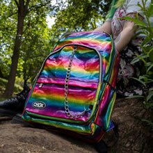 Load image into Gallery viewer, The CHOK rainbow holographic vegan shown next to a green haired model sat on a tree trunk. The bag has a rainbow melt holographic print with a detachable silver chain and CHOK logo. The bag is facing forward to highlight the front rainbow zip pockets, the main double rainbow zip compartment, two side elasticated pockets with a detachable decorative silver chain.