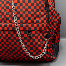 Load image into Gallery viewer, Close up of the Red Checkerboard Backpack sat on a grey background. The vegan friendly bag is facing forward to highlight the red and black check print, two front zip pockets, two elasticated side pockets, main top double zip pocket and silver draping decorative chain.