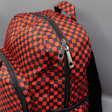 Load image into Gallery viewer, Close up of the Red Checkerboard Backpack sat on a grey background. The vegan friendly bag is facing forward to highlight the red and black check print, two front zip pockets, two elasticated side pockets, main top double zip pocket and silver draping decorative chain.