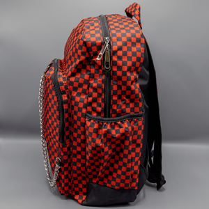 The Red Checkerboard Backpack sat on a grey background. The vegan friendly bag is facing left to highlight the red and black check print, two front zip pockets, two elasticated side pockets, main top double zip pocket and silver draping decorative chain.