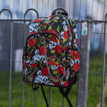 Load image into Gallery viewer, The skulls and roses vegan backpack hanging on a skatepark railing. The bag is facing forward to highlight the skulls and roses all over print, silver chain, two zip front pockets and two side elastic pockets.