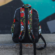 Load image into Gallery viewer, Skulls and roses vegan backpack sat on a skatepark bench in front of a graffiti wall. The bag is facing away to highlight the skulls and roses printed padded adjustable shoulder straps, two elasticated side pockets and plain black back.
