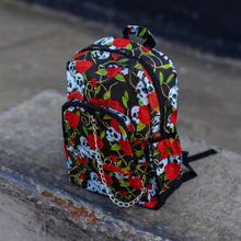 Load image into Gallery viewer, The skulls and roses vegan backpack sat on a skatepark bench. The bag is facing forward angled to the left to highlight the skulls and roses all over print, silver chain, two zip front pockets and two side elastic pockets.