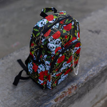 Load image into Gallery viewer, The skulls and roses vegan backpack sat on a skatepark bench. The bag is facing forward angled to the right to highlight the skulls and roses all over print, silver chain, two zip front pockets and two side elastic pockets.