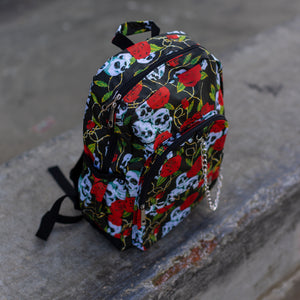 The skulls and roses vegan backpack sat on a skatepark bench. The bag is facing forward angled to the right to highlight the skulls and roses all over print, silver chain, two zip front pockets and two side elastic pockets.
