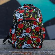 Load image into Gallery viewer, Skulls and roses vegan backpack sat on a skatepark bench in front of a graffiti wall. The bag is facing forward to highlight the skulls and roses all over print, silver chain, two zip front pockets and two side elastic pockets.
