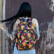 Load image into Gallery viewer, Model standing in front of a brick wall wearing the floral gold skull nylon chain vegan backpack. The bag is facing the camera to highlight the flowers and skulls print, two zipped pockets, two elastic side pockets and detachable decorative silver chain.